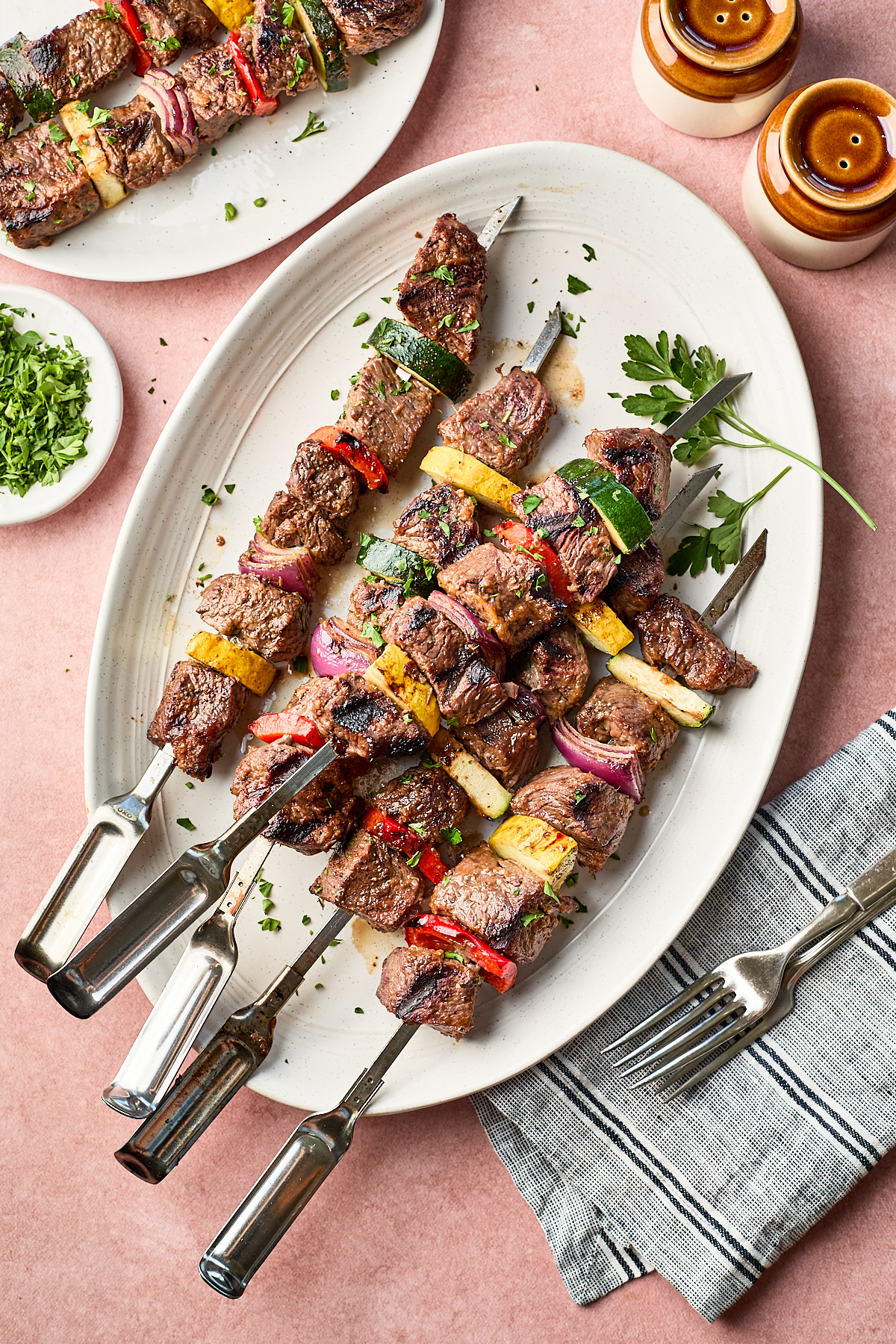 Grilled beef kebabs with colorful bell peppers, zucchini, and red onions are arranged on a white oval platter, garnished with herbs. Two forks, napkins, salt and pepper shakers, and a dish of chopped herbs are nearby.