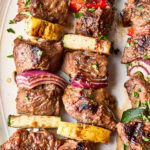 Grilled beef kebabs on skewers with pieces of red onion, zucchini, yellow squash, and red bell pepper, garnished with chopped herbs on a white plate.