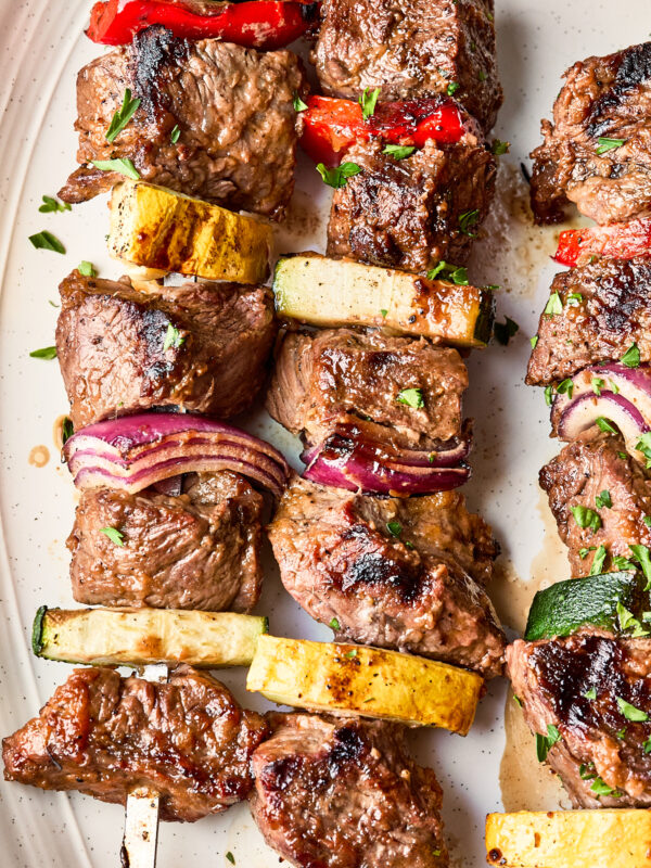 Grilled beef kebabs on skewers with pieces of red onion, zucchini, yellow squash, and red bell pepper, garnished with chopped herbs on a white plate.