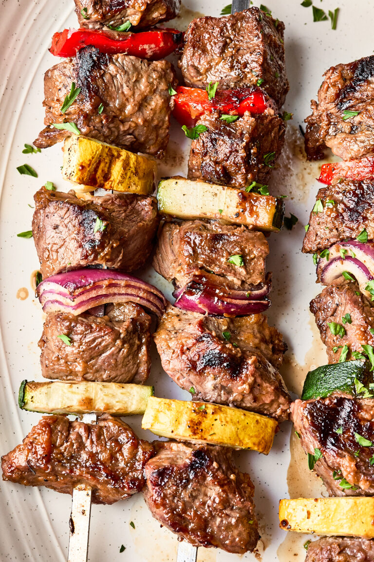 Grilled beef kebabs on skewers with pieces of red onion, zucchini, yellow squash, and red bell pepper, garnished with chopped herbs on a white plate.