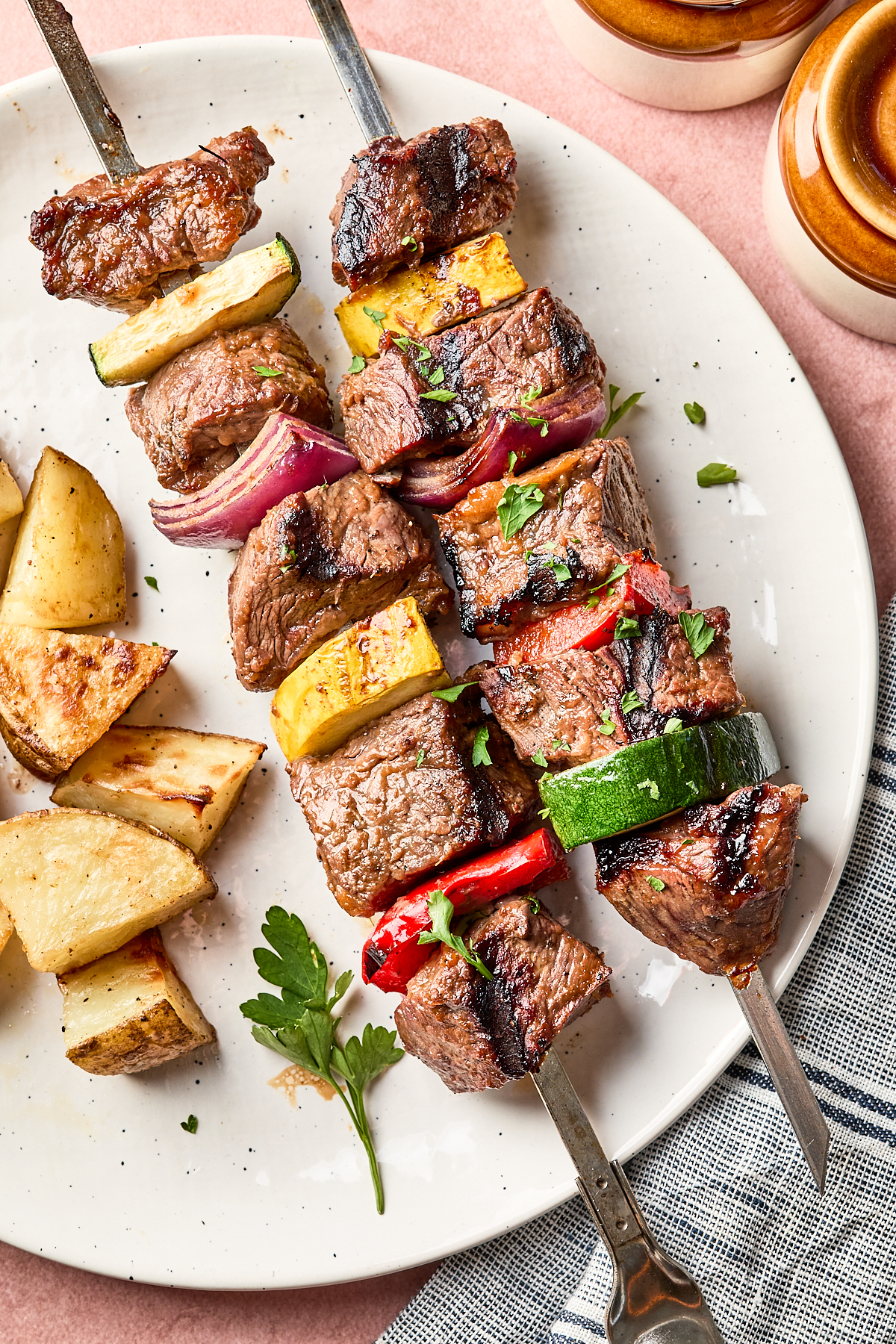 Two metal skewers with grilled beef cubes, zucchini, red onion, and red bell pepper on a white plate, served with roasted potato wedges and garnished with fresh parsley.