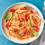 A plate of spaghetti with tomato sauce, garnished with fresh basil leaves, sits on a blue surface next to a striped napkin and a fork resting on the plate.