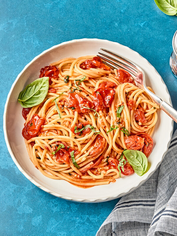 A plate of spaghetti with tomato sauce, garnished with fresh basil leaves, sits on a blue surface next to a striped napkin and a fork resting on the plate.