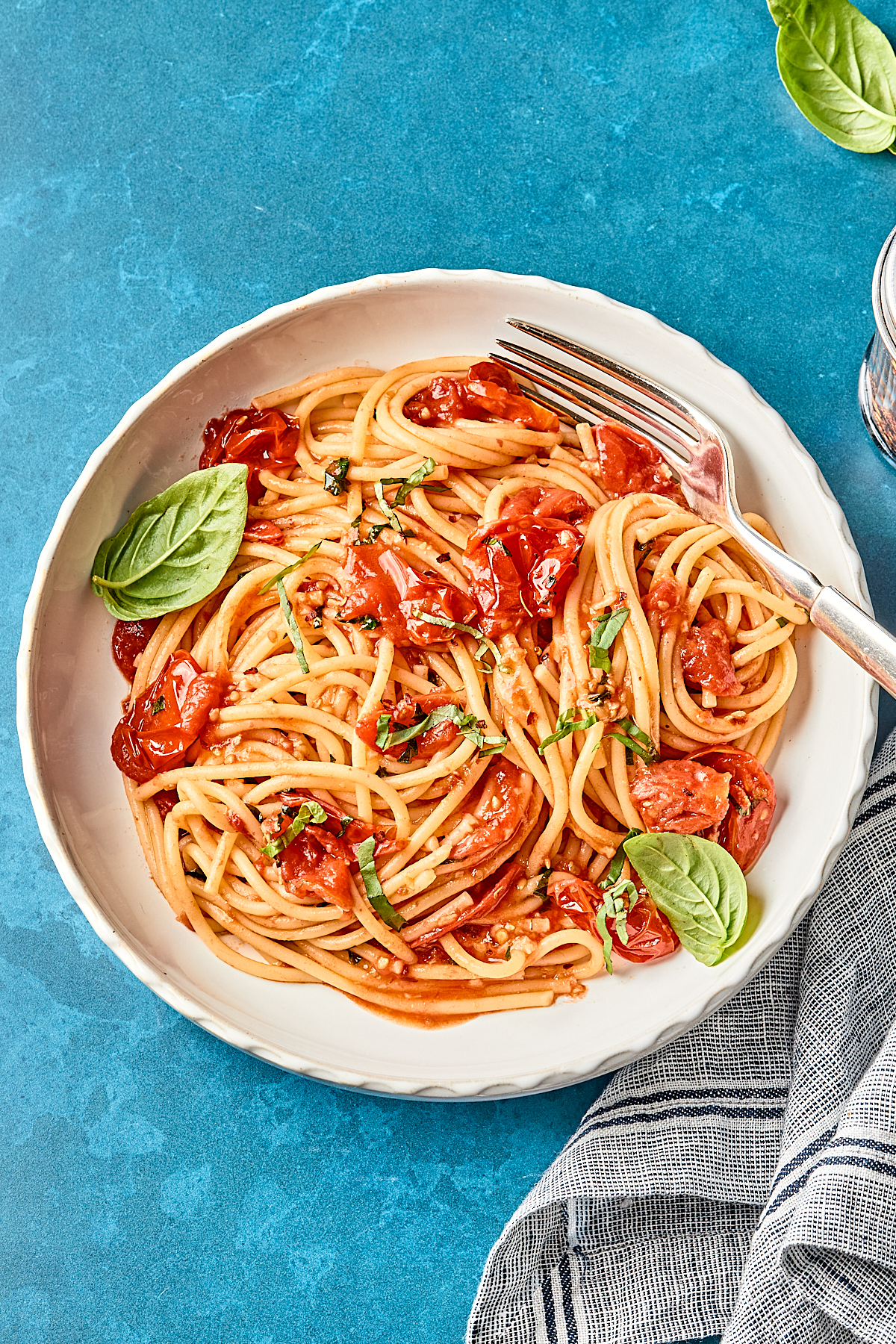 A plate of spaghetti with tomato sauce, garnished with fresh basil leaves, sits on a blue surface next to a striped napkin and a fork resting on the plate.