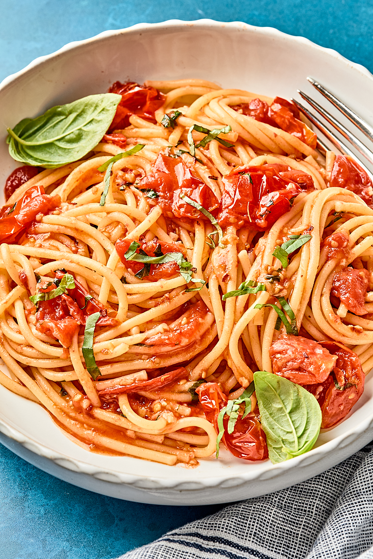 A white bowl of spaghetti topped with roasted cherry tomatoes, fresh basil leaves, and a tomato-based sauce, placed on a blue surface with a fork on the side and a striped napkin partially visible.