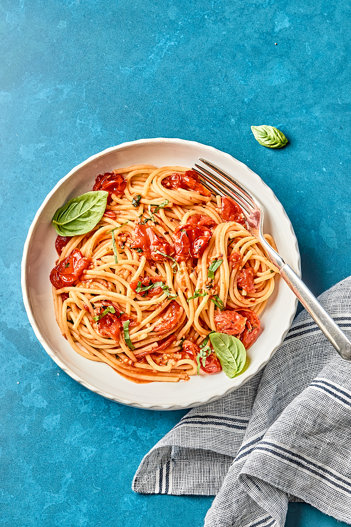 A white plate of spaghetti with tomato sauce, garnished with basil leaves, sits on a blue surface beside a striped cloth napkin and a fork.