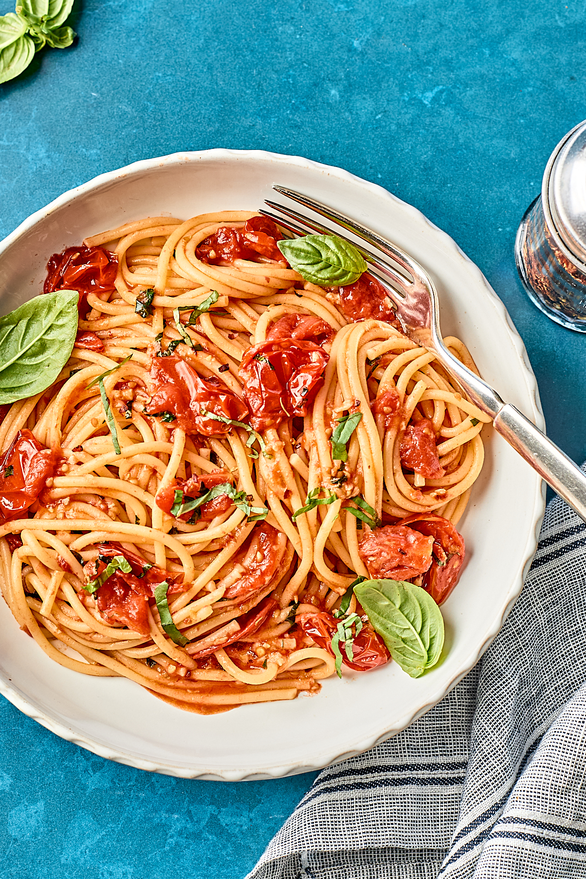 A plate of spaghetti with roasted cherry tomatoes, fresh basil leaves, and a fork on the side, set on a blue table next to a striped cloth napkin.