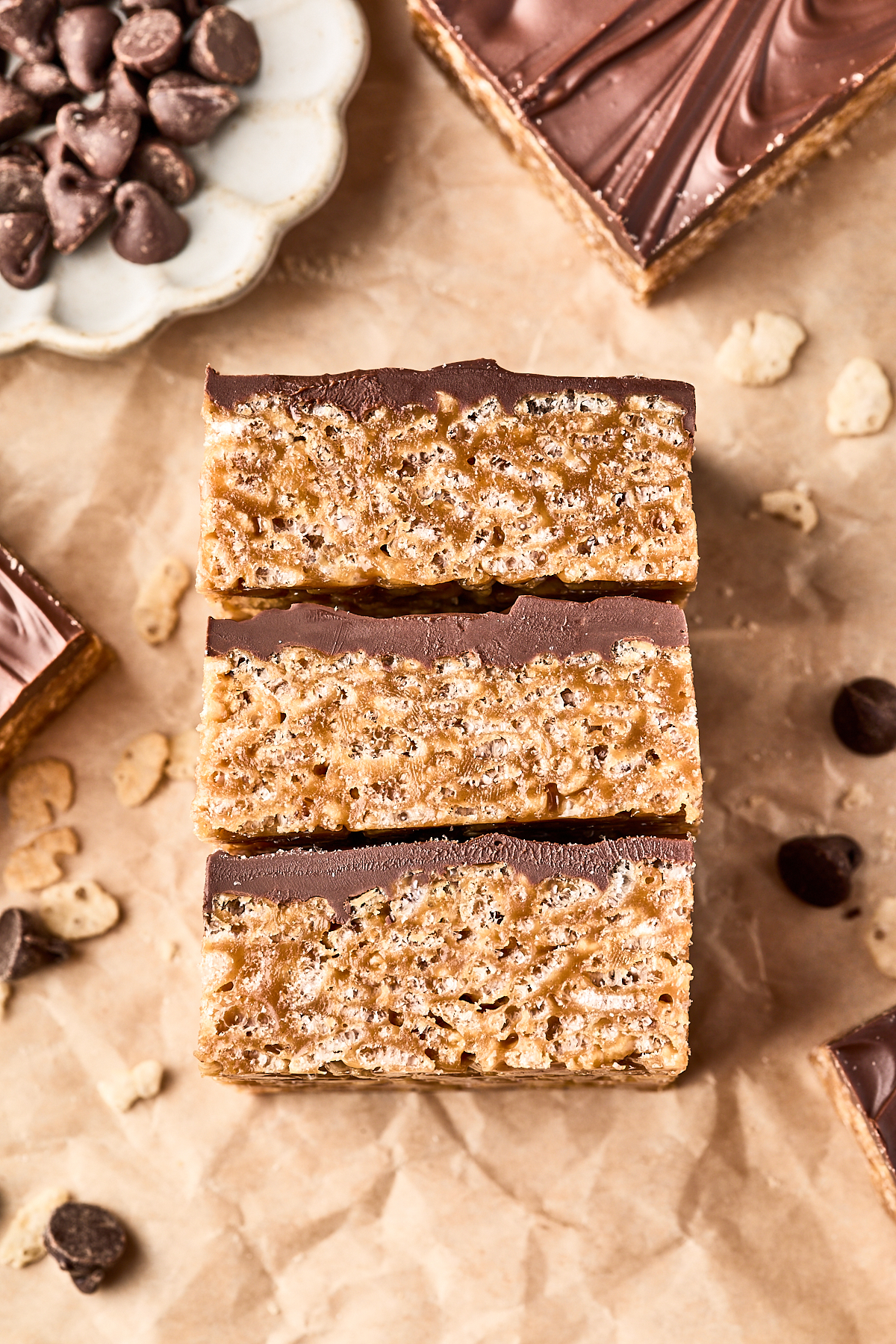 Three chocolate-topped Rice Krispies treats are stacked on parchment paper, with chocolate chips and a plate of more chips nearby. The treats have a crispy, layered texture.