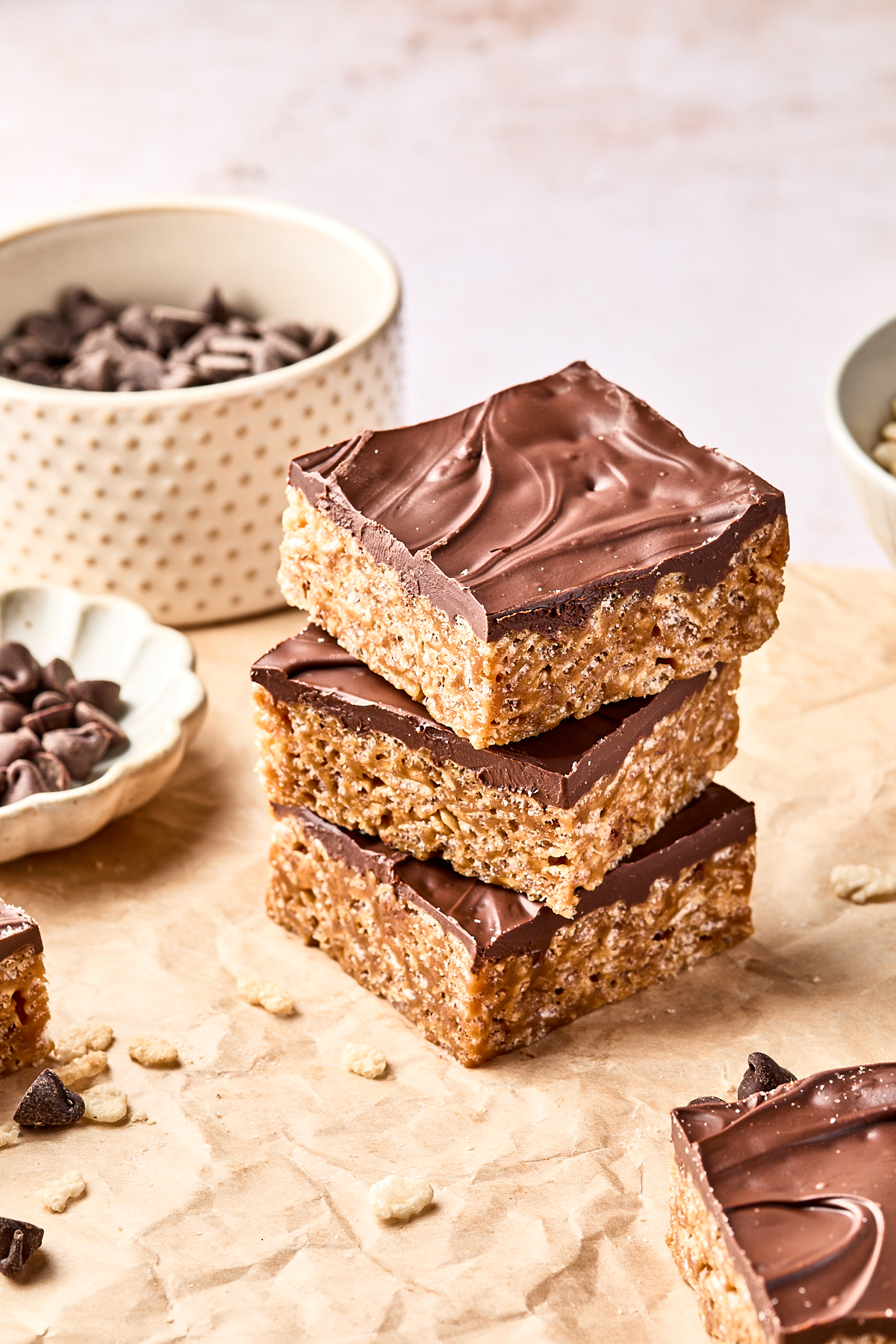 Three chocolate-topped rice crispy treats are stacked on parchment paper, surrounded by bowls of chocolate chips and scattered treat crumbs.