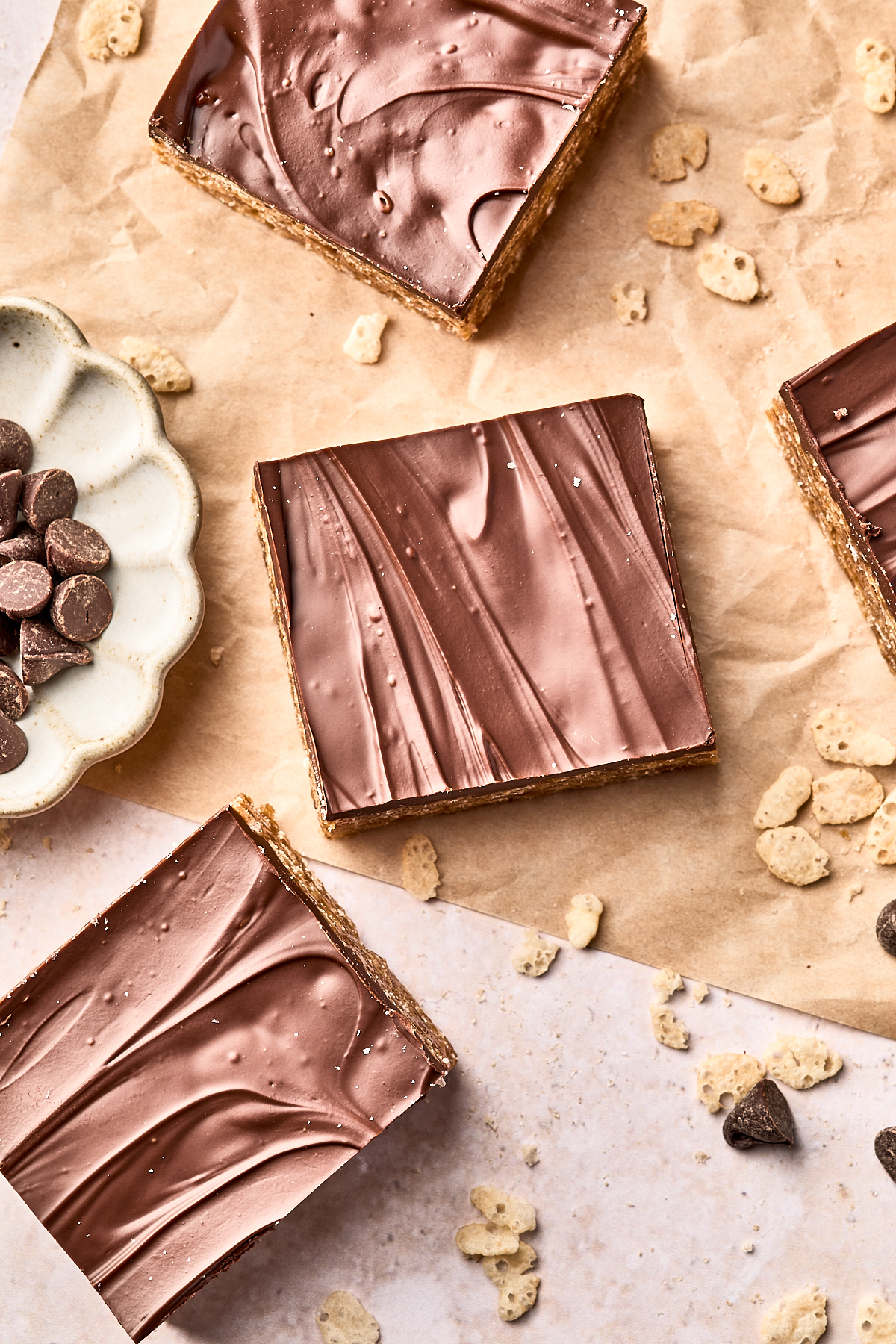 Four chocolate-topped dessert bars rest on parchment paper, surrounded by chocolate chips and scattered cereal pieces. The bars have a shiny, swirled chocolate layer on top.