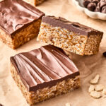 Three chocolate-topped rice crispy treats are stacked on parchment paper, with more treats and a small bowl of chocolate chips in the background. Some cereal crumbs are scattered nearby.