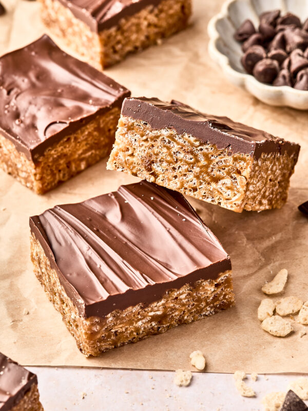Three chocolate-topped rice crispy treats are stacked on parchment paper, with more treats and a small bowl of chocolate chips in the background. Some cereal crumbs are scattered nearby.
