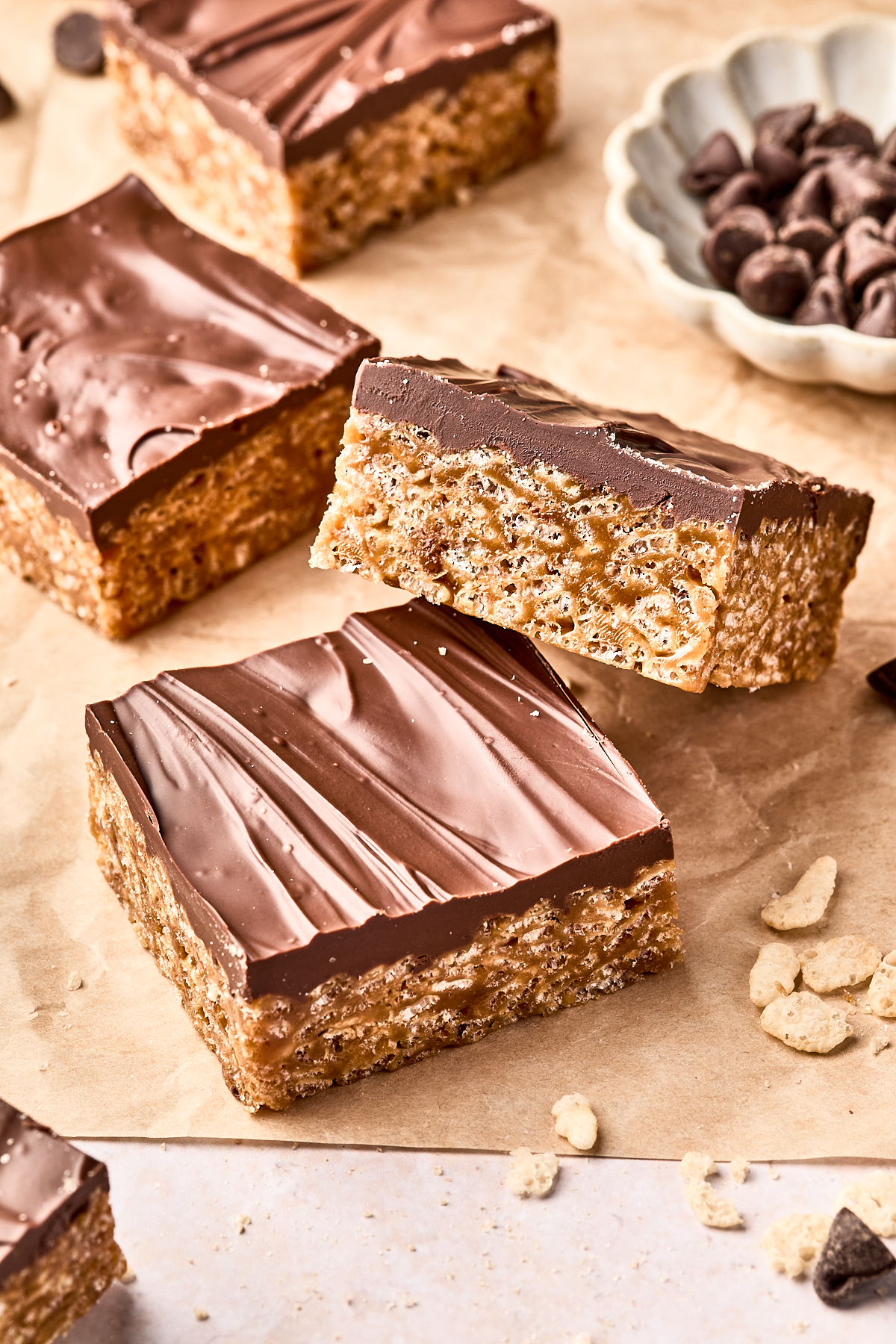 Three chocolate-topped rice crispy treats are stacked on parchment paper, with more treats and a small bowl of chocolate chips in the background. Some cereal crumbs are scattered nearby.