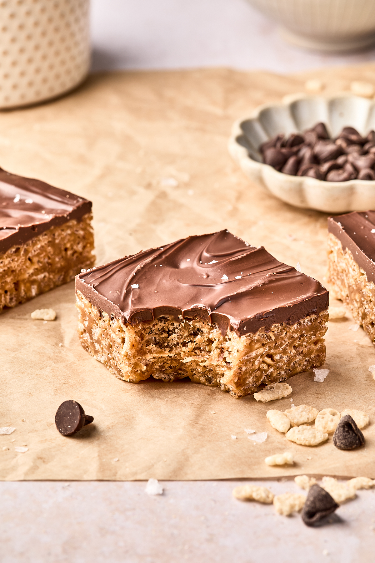 A chocolate-covered rice crispy treat with a bite taken out, surrounded by chocolate chips and cereal pieces on parchment paper.