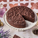 A chocolate crumb cake with chocolate chips sits on a white cake stand. A single slice is cut out and placed on a plate with a fork. Decorative items and flowers surround the cake.
