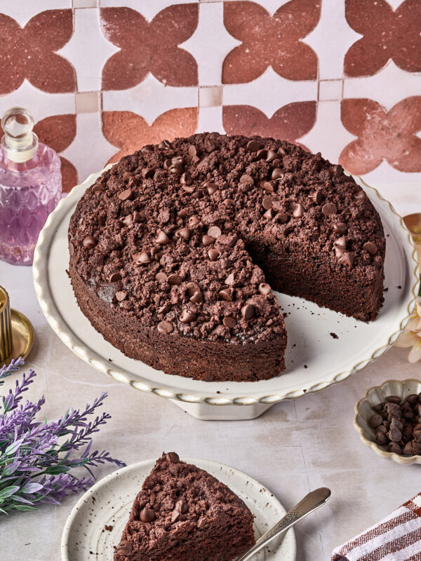 A chocolate crumb cake with chocolate chips sits on a white cake stand. A single slice is cut out and placed on a plate with a fork. Decorative items and flowers surround the cake.