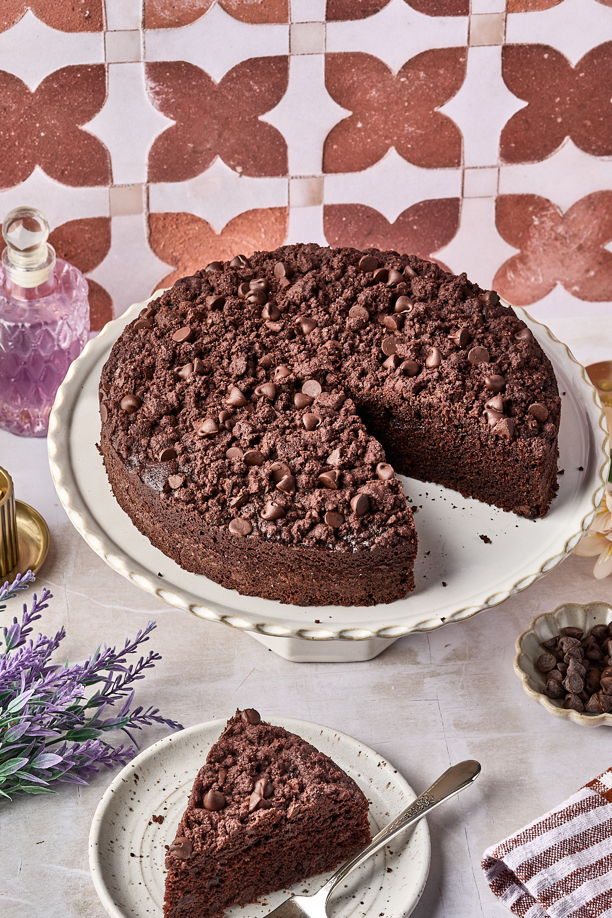 A chocolate crumb cake with chocolate chips sits on a white cake stand. A single slice is cut out and placed on a plate with a fork. Decorative items and flowers surround the cake.