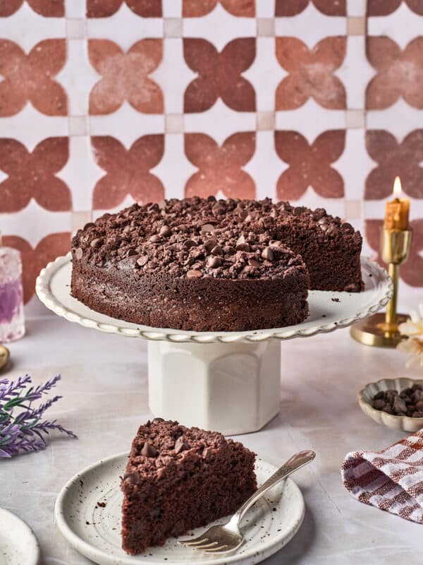 A round chocolate cake with chocolate chips sits on a cake stand, with a slice on a plate in front and a fork beside it. The background features a pink patterned tile wall and a lit candle.