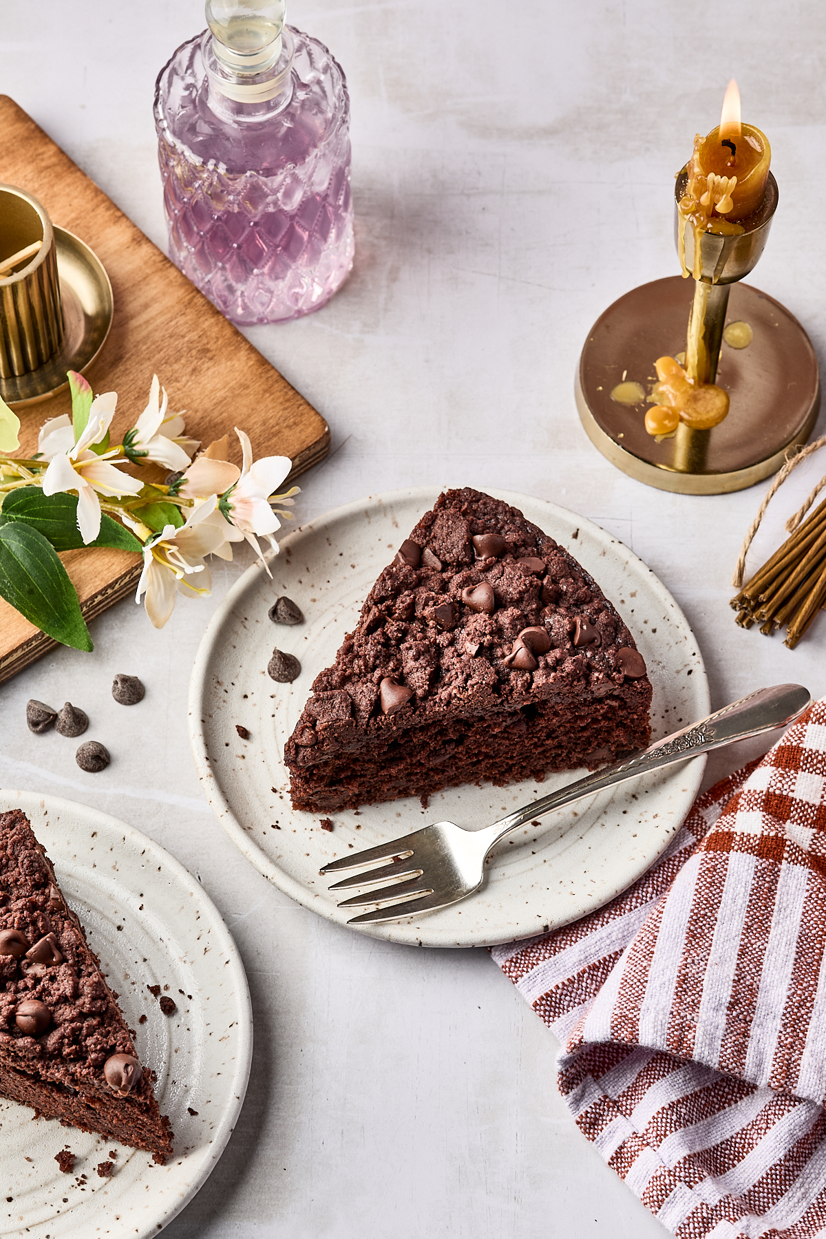 A slice of chocolate cake with chocolate chips on a plate with a fork, beside a candle, a glass bottle, flowers, and a striped napkin on a light surface.
