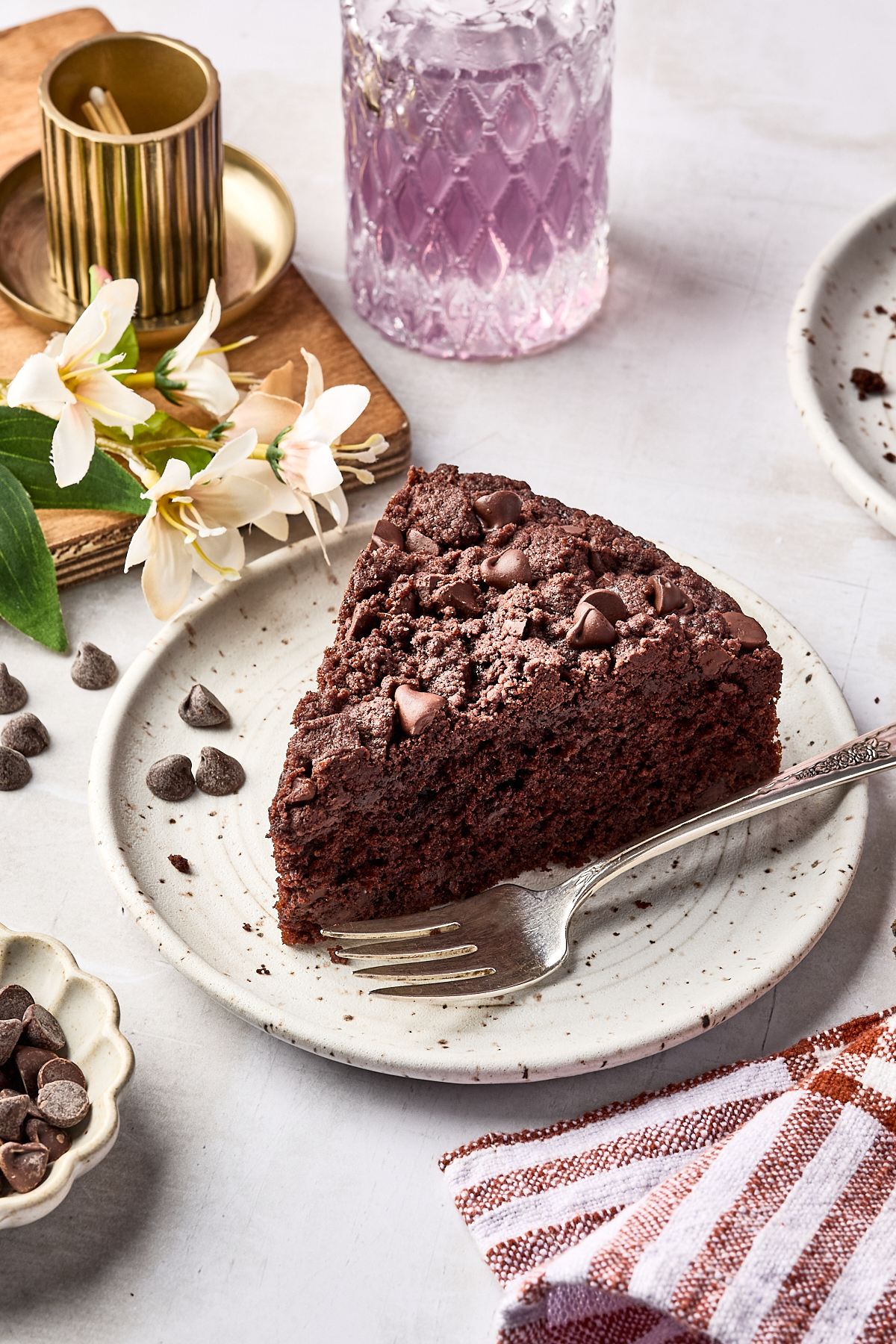 A slice of chocolate cake with chocolate chips on a plate, a fork beside it, flowers, a glass of purple drink, and scattered chocolate chips on the table.