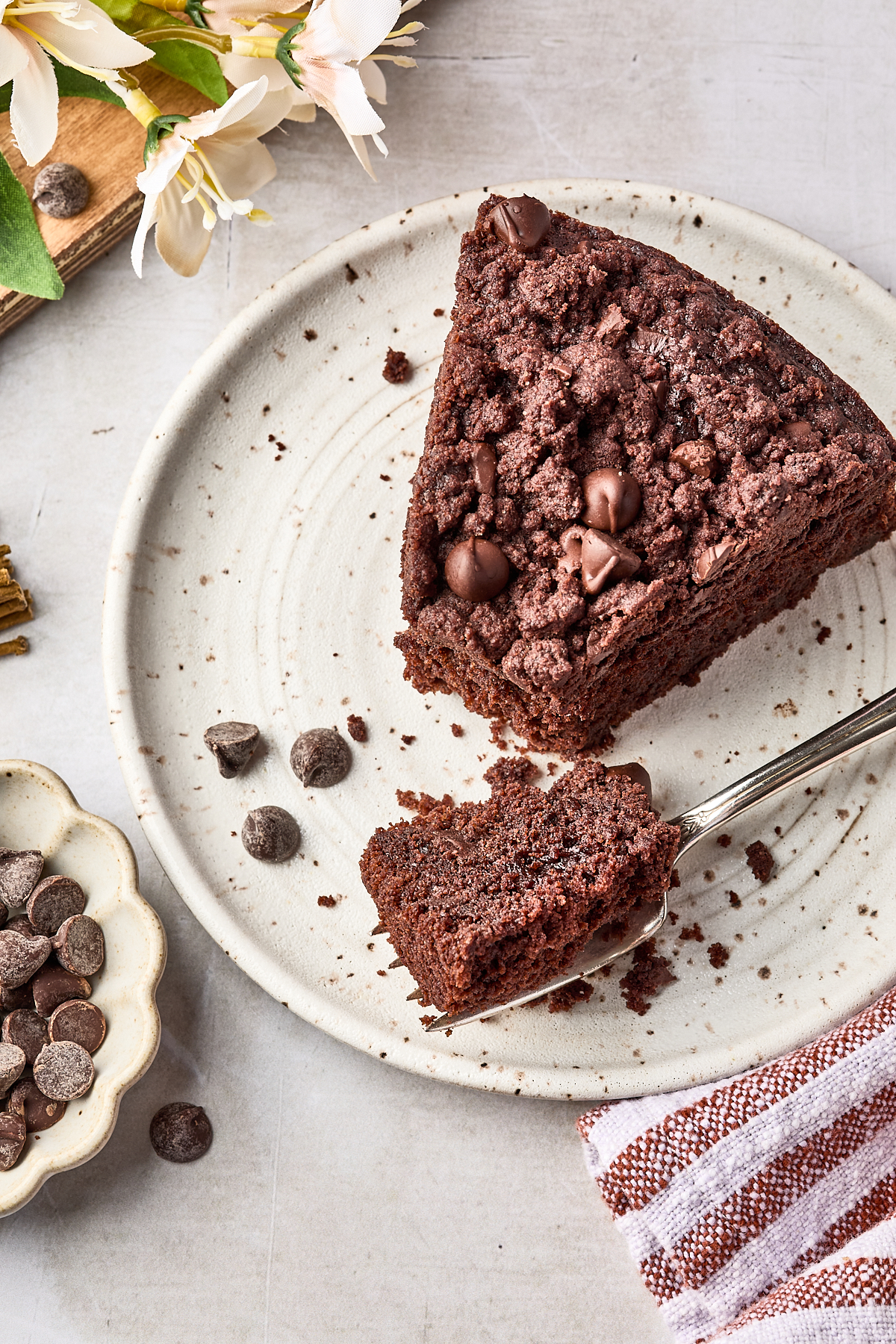 A slice of chocolate chip crumb cake on a ceramic plate, with a fork cutting a bite. Chocolate chips are scattered nearby, and a striped towel and flowers are visible at the edge.