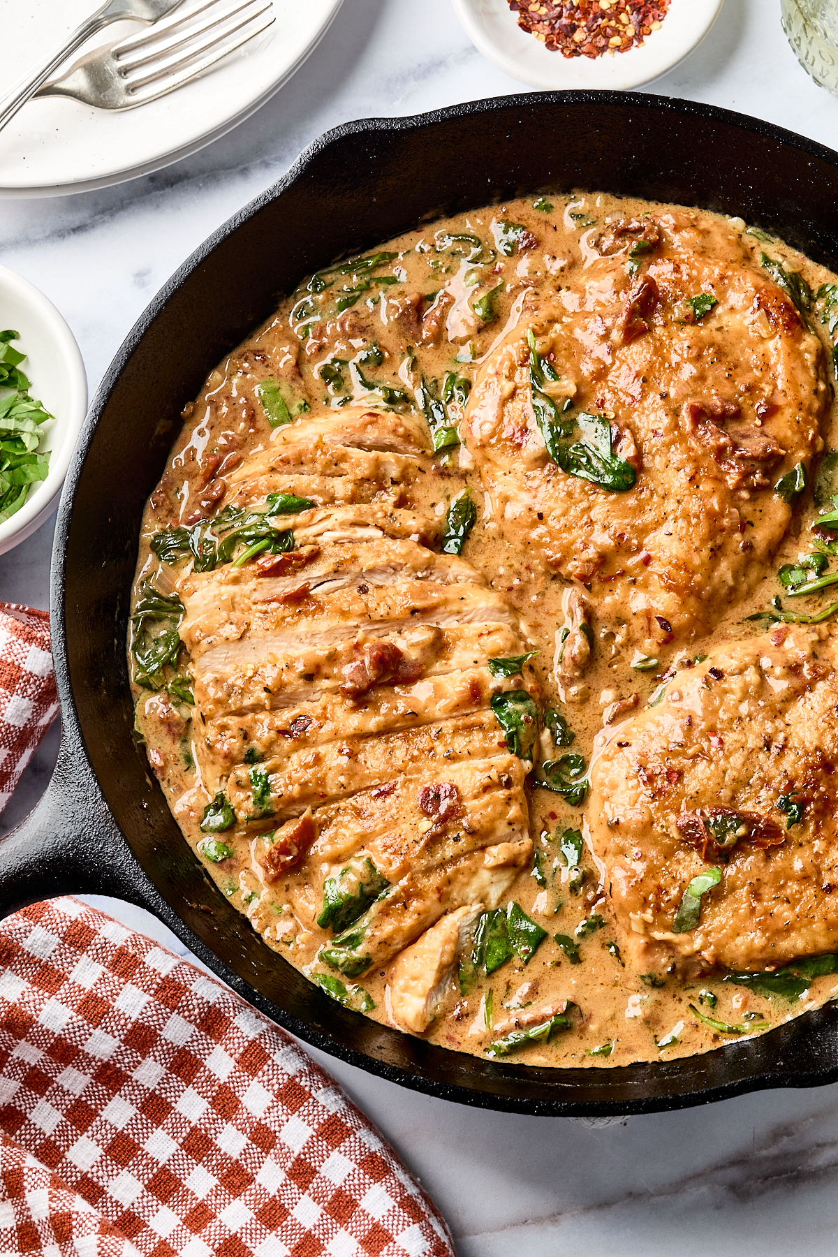 Three cooked chicken breasts in a creamy sauce with spinach in a black skillet, next to a small bowl of chopped herbs and a red-checked towel.