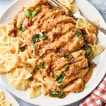 Plate of bowtie pasta topped with sliced chicken in a creamy sauce with sun-dried tomatoes and spinach. A fork rests on the plate, and a red-checkered napkin is nearby.