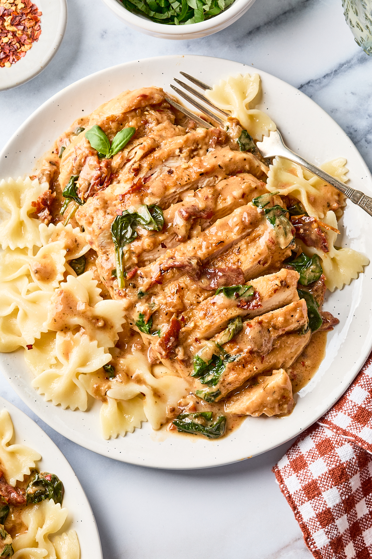 Plate of bowtie pasta topped with sliced chicken in a creamy sauce with sun-dried tomatoes and spinach. A fork rests on the plate, and a red-checkered napkin is nearby.