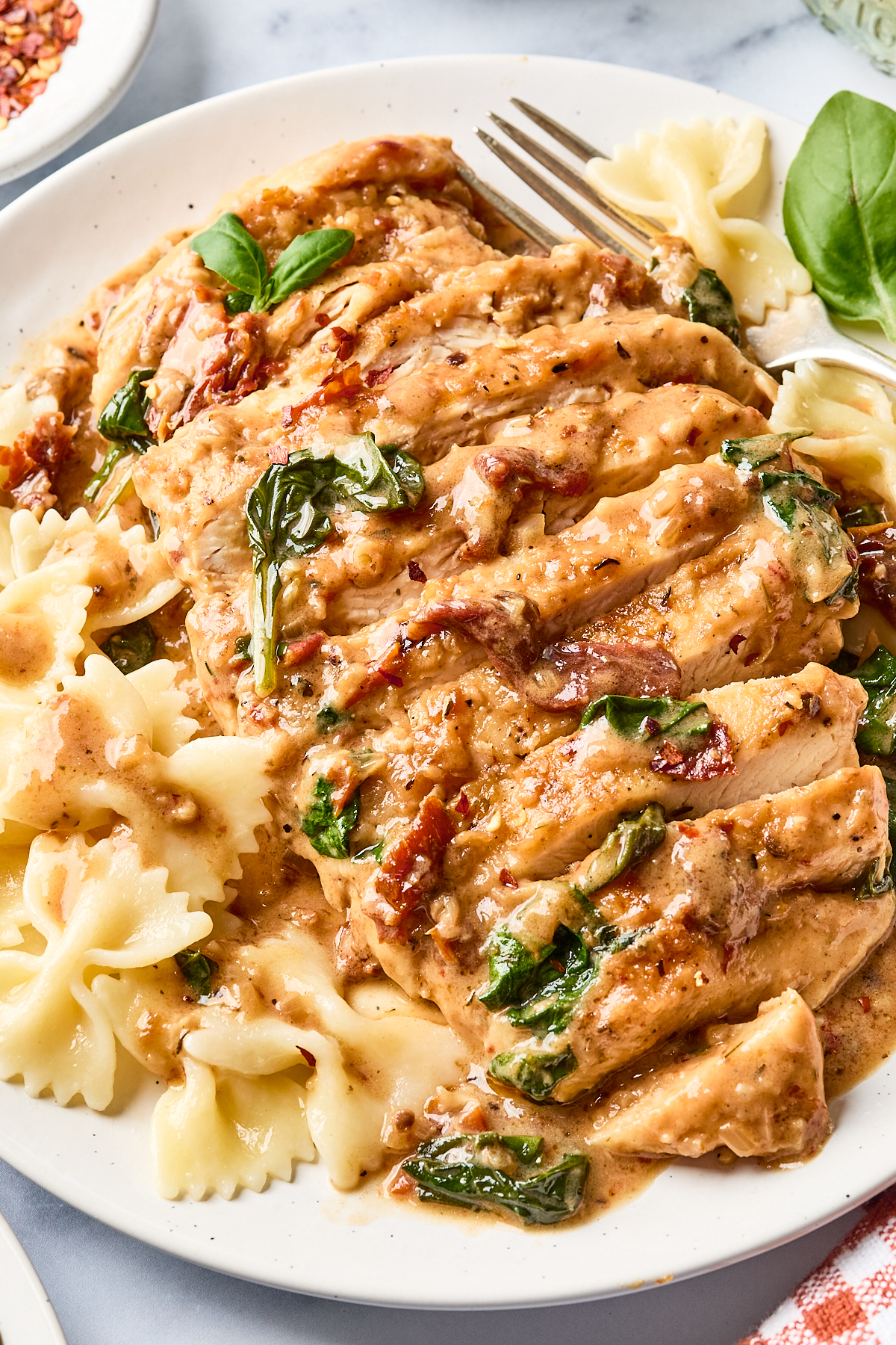 Sliced chicken breast in creamy sauce with spinach and sun-dried tomatoes, served with bowtie pasta on a white plate. A fork and fresh basil garnish are visible beside the food.