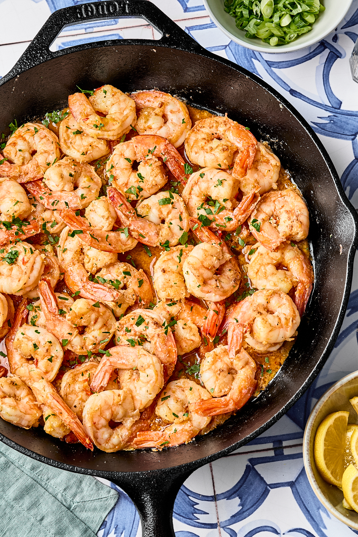 Shrimp cooked in a skillet with sauce, garnished with chopped herbs. A bowl of sliced green onions and a bowl of lemon wedges are nearby on a patterned blue and white surface.
