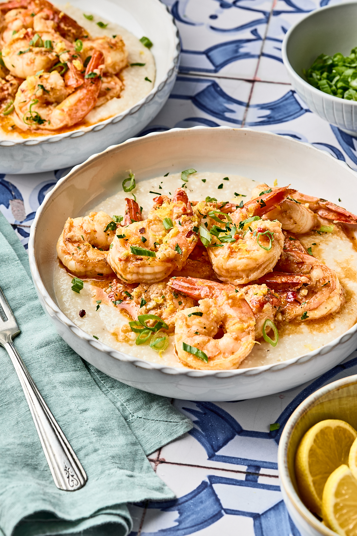A bowl of creamy grits topped with sautéed shrimp, garnished with chopped green onions, sits on a patterned tile surface next to a fork, lemon slices, and a bowl of more green onions.