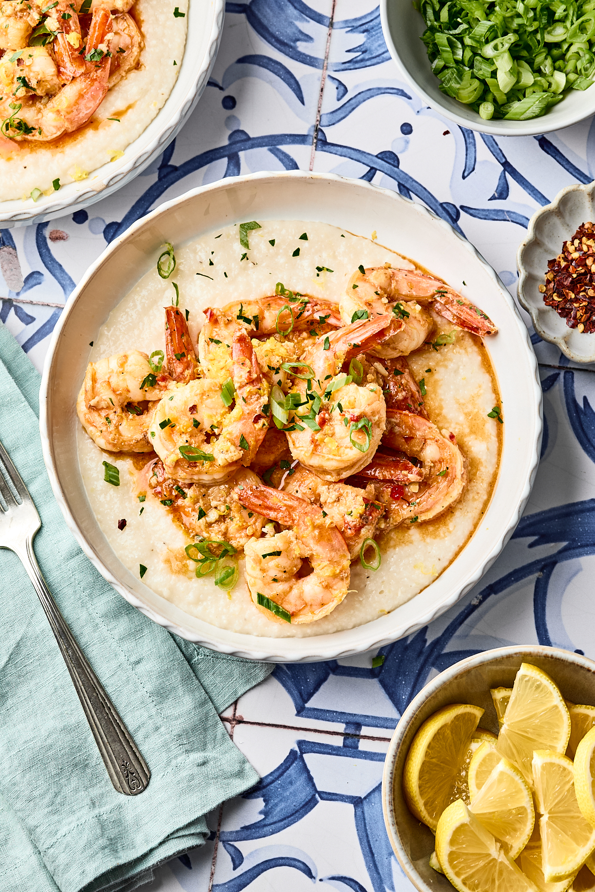 A bowl of creamy grits topped with cooked shrimp, garnished with chopped green onions, sits on a patterned blue surface. Lemon wedges, red pepper flakes, and more green onions are on the side.