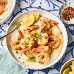 A bowl of shrimp served over creamy grits, garnished with green onions and lemon wedges, with small bowls of sliced green onions, red pepper flakes, and lemon wedges nearby on a patterned blue and white table.