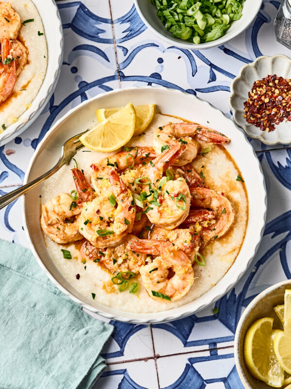 A bowl of shrimp served over creamy grits, garnished with green onions and lemon wedges, with small bowls of sliced green onions, red pepper flakes, and lemon wedges nearby on a patterned blue and white table.
