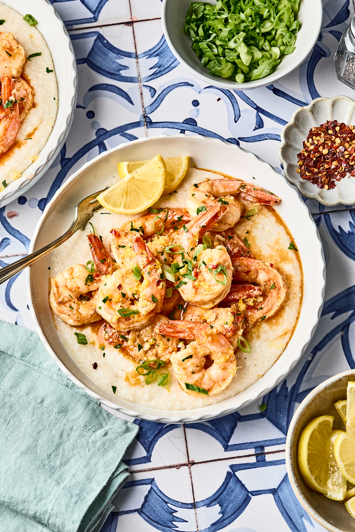 A bowl of shrimp served over creamy grits, garnished with green onions and lemon wedges, with small bowls of sliced green onions, red pepper flakes, and lemon wedges nearby on a patterned blue and white table.