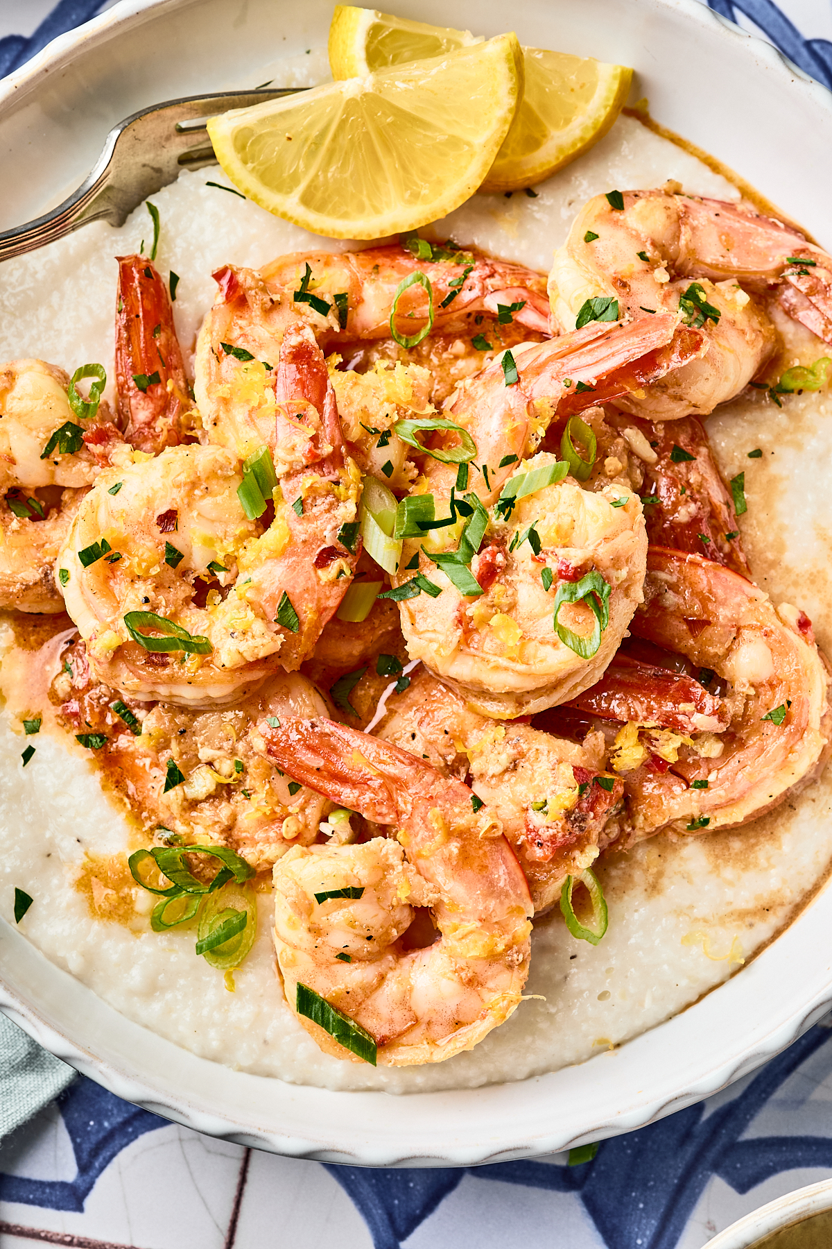 A plate of shrimp in a garlic sauce served over creamy grits, garnished with chopped green onions and lemon wedges.