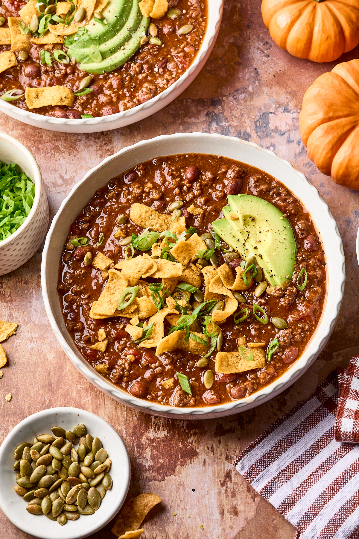 A bowl of chili topped with avocado slices, corn chips, green onions, and seeds; a small bowl of seeds and a cloth napkin are nearby, with another bowl of chili and two pumpkins in the background.