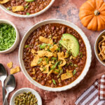Two bowls of chili topped with avocado slices, corn chips, and green onions. Surrounding items include sliced lettuce, pumpkin seeds, spoons, two small pumpkins, and a striped towel on a rustic surface.