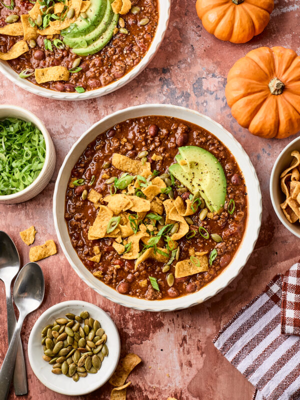 Two bowls of chili topped with avocado slices, corn chips, and green onions. Surrounding items include sliced lettuce, pumpkin seeds, spoons, two small pumpkins, and a striped towel on a rustic surface.