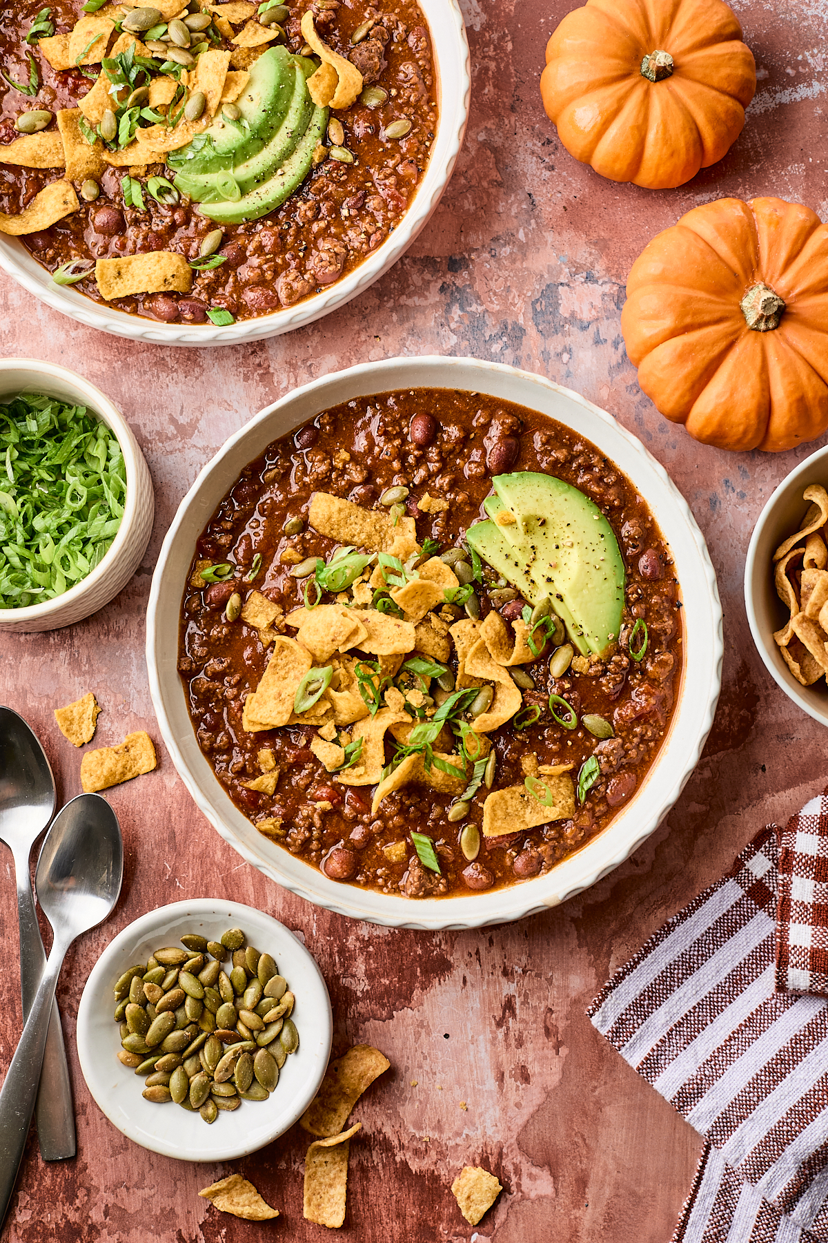 Two bowls of chili topped with avocado slices, corn chips, and green onions. Surrounding items include sliced lettuce, pumpkin seeds, spoons, two small pumpkins, and a striped towel on a rustic surface.