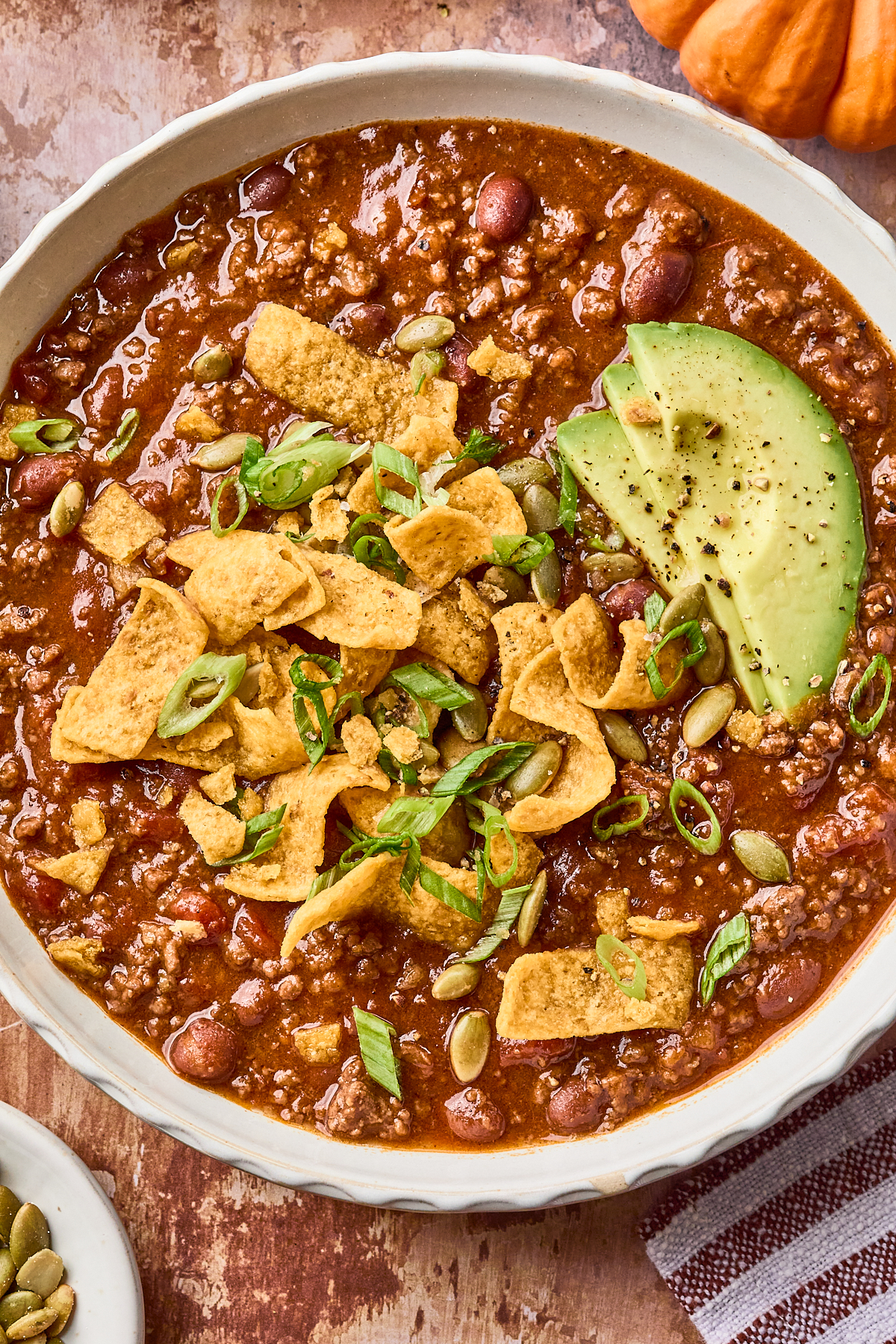A bowl of chili topped with corn chips, pumpkin seeds, sliced green onions, and avocado slices, set on a brown surface with a small pumpkin and a striped napkin nearby.