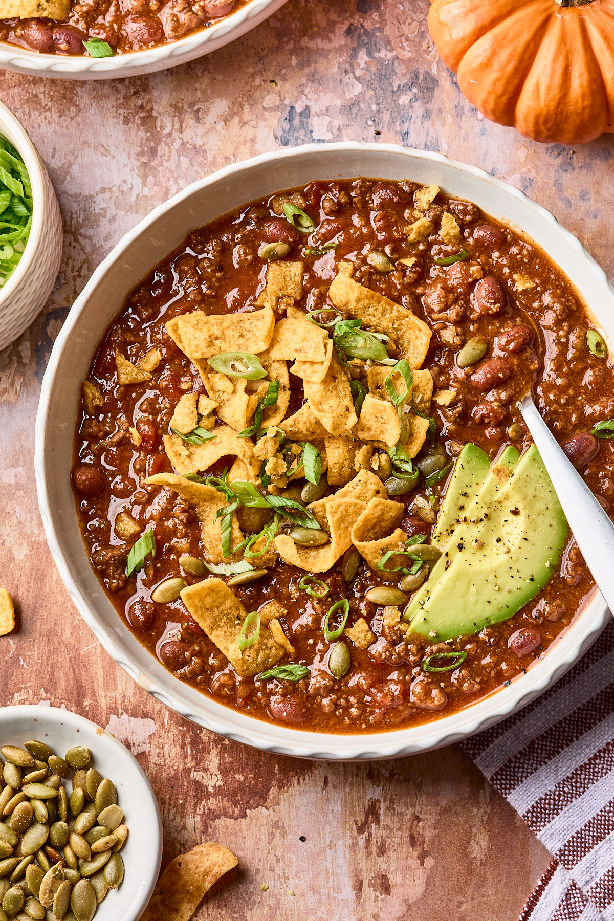 A bowl of chili topped with corn chips, sliced avocado, green onions, and pumpkin seeds sits on a rustic surface, surrounded by small bowls of ingredients and a mini pumpkin.