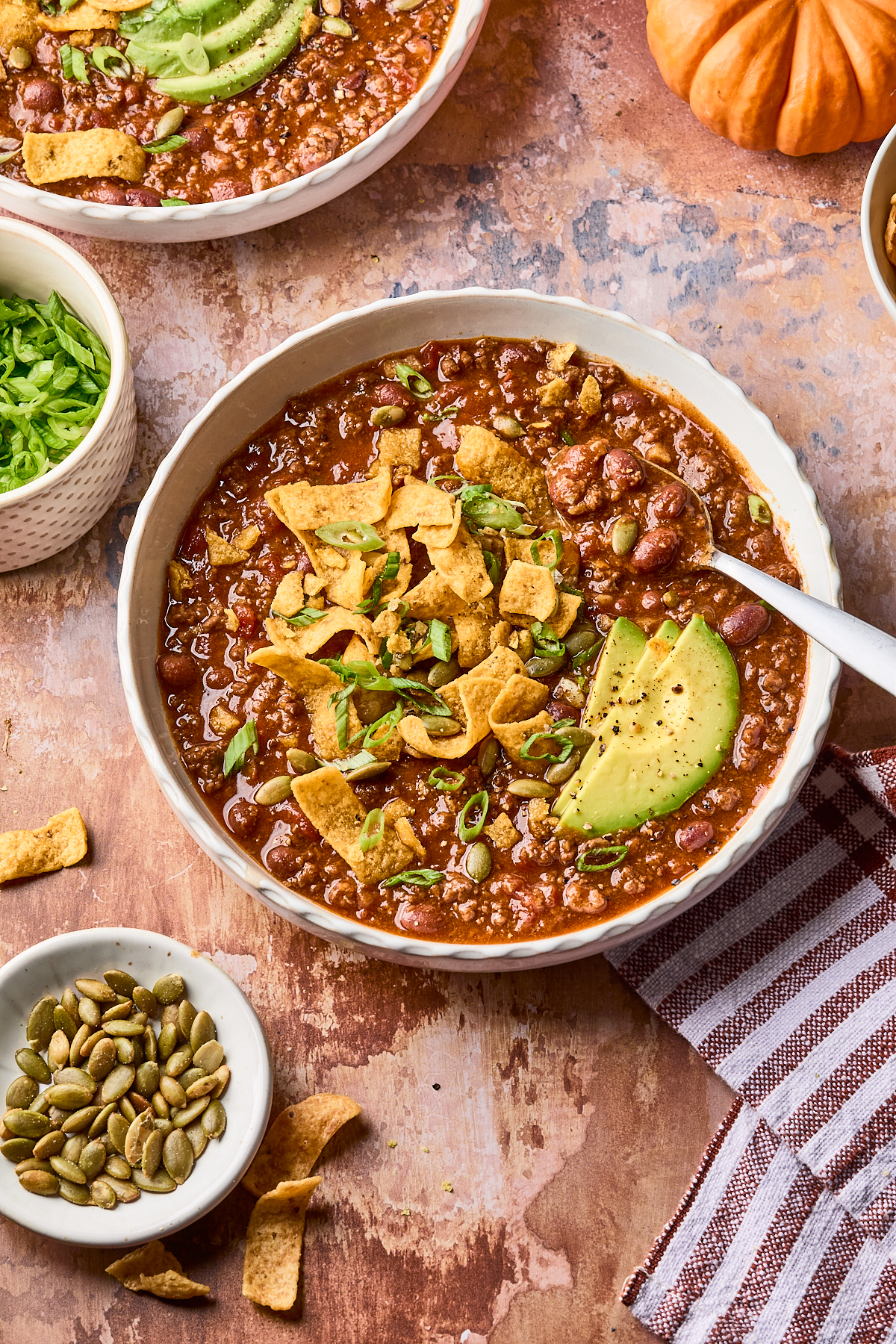 A bowl of chili topped with corn chips, avocado slices, and green onions, next to a bowl of pumpkin seeds, a bowl of chopped lettuce, and a striped napkin on a rustic surface.