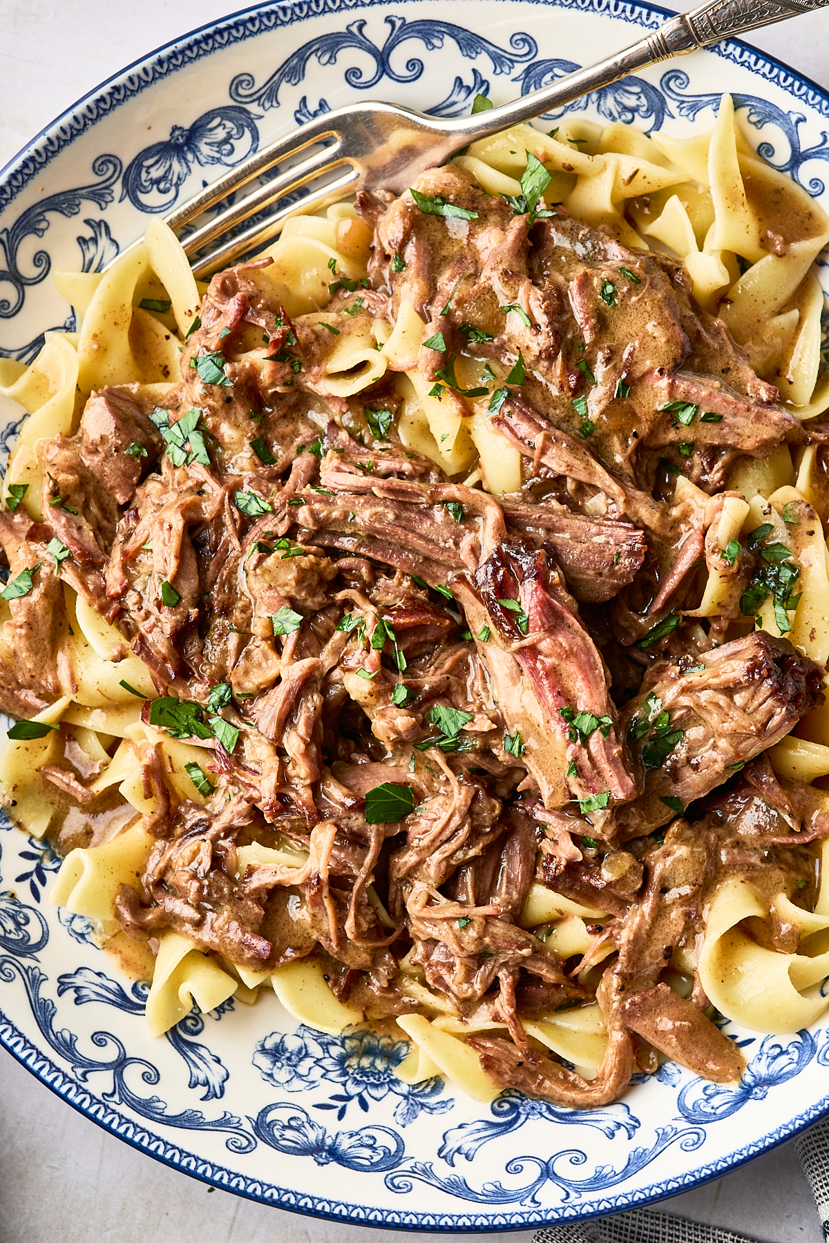 Slow-cooked shredded beef in a savory gravy served over egg noodles in a blue and white patterned bowl, with a fork resting on the side.