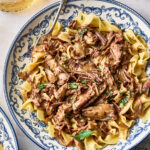 Beef stroganoff with shredded meat and wide egg noodles served in a blue and white patterned bowl with a silver fork.