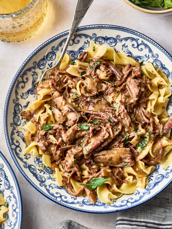 Beef stroganoff with shredded meat and wide egg noodles served in a blue and white patterned bowl with a silver fork.