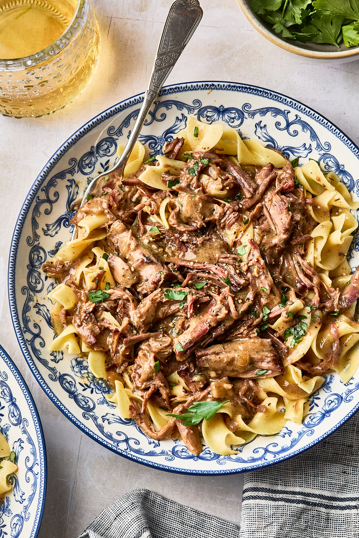 Beef stroganoff with shredded meat and wide egg noodles served in a blue and white patterned bowl with a silver fork.