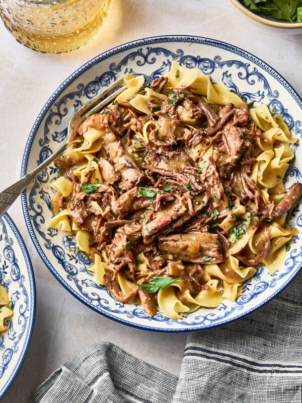 Beef stroganoff with shredded meat served over egg noodles in a blue and white patterned bowl, with a fork resting on the side.