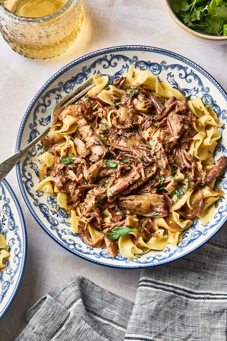 Beef stroganoff with shredded meat served over egg noodles in a blue and white patterned bowl, with a fork resting on the side.